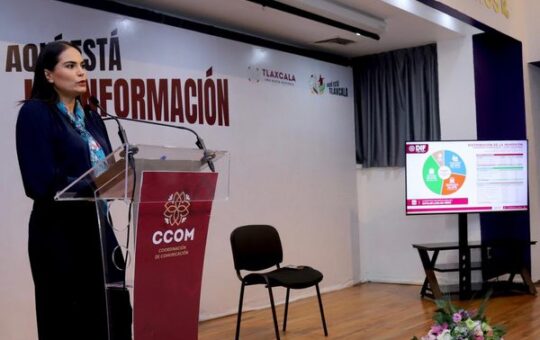 Mujer habla ante micrófonos en un podio de la CCOM de Tlaxcala; al fondo, una pantalla con una gráfica del DIF