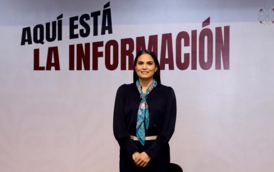 Mujer con blusa azul marino y pañuelo turquesa posa de pie ante una pared con el letrero Aquí está la información