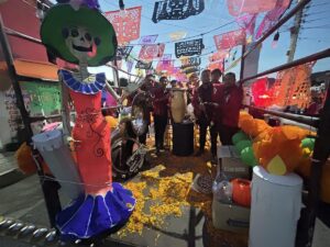 Banda de músicos con camisas rojas posa junto a una catrina de cartón, flores de cempasúchil y papel picado en una calle...