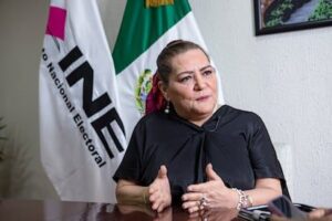 Mujer de blusa negra gesticula mientras habla, sentada en una oficina junto a banderas del INE y de México