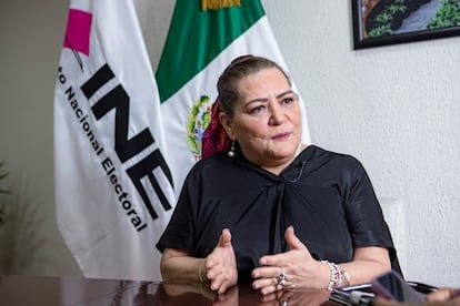 Mujer de blusa negra gesticula mientras habla, sentada en una oficina junto a banderas del INE y de México
