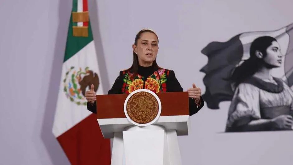 Mujer con blusa bordada de flores habla en un podio con el escudo nacional, junto a una bandera de México