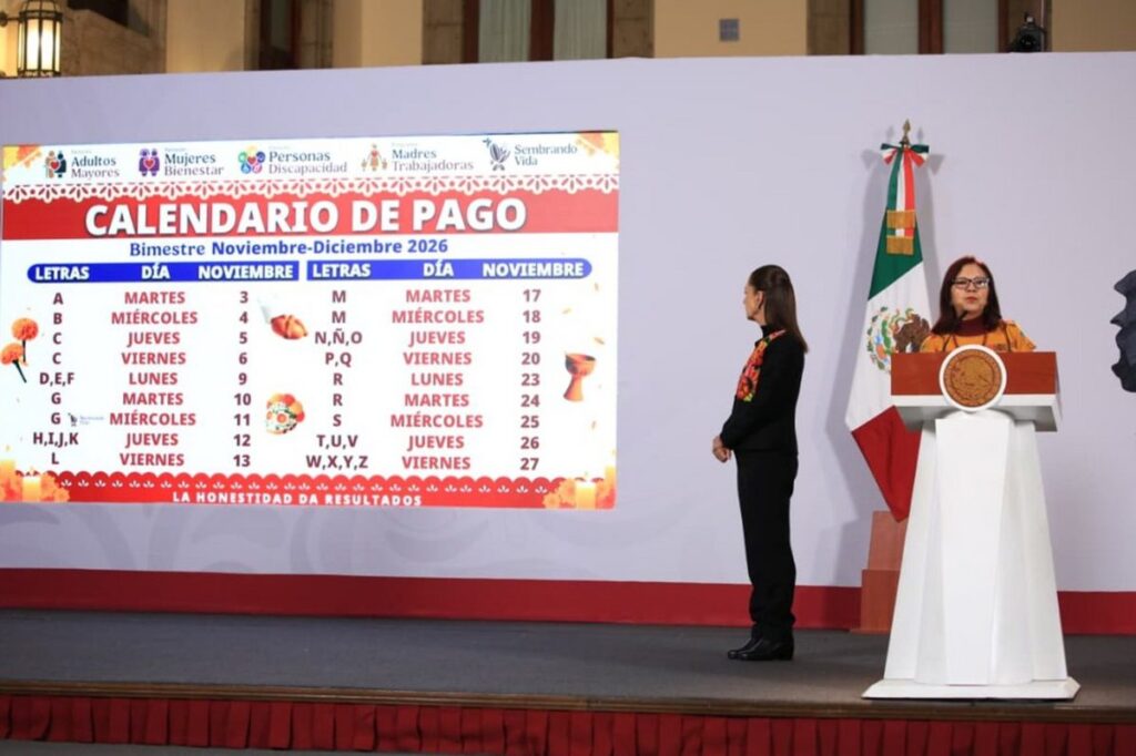 Una mujer habla en un podio y otra, de pie, mira la pantalla con el calendario de pago de noviembre de Bienestar