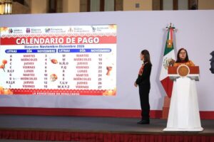 Una mujer habla en un podio y otra, de pie, mira la pantalla con el calendario de pago de noviembre de Bienestar