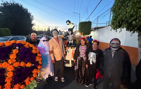 Grupo de personas disfrazadas de calaveras y fantasma posa junto a un arco de cempasúchil y un esqueleto decorativo en...