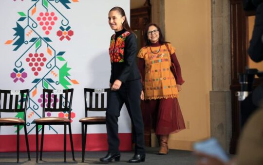 Dos mujeres con prendas bordadas caminan sonrientes por un escenario con sillas vacías y un fondo de motivos florales