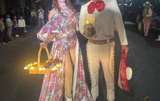 Pareja caracterizada de catrina y charro calavera posa de noche en el desfile de Panotla; ella lleva una canasta con velas