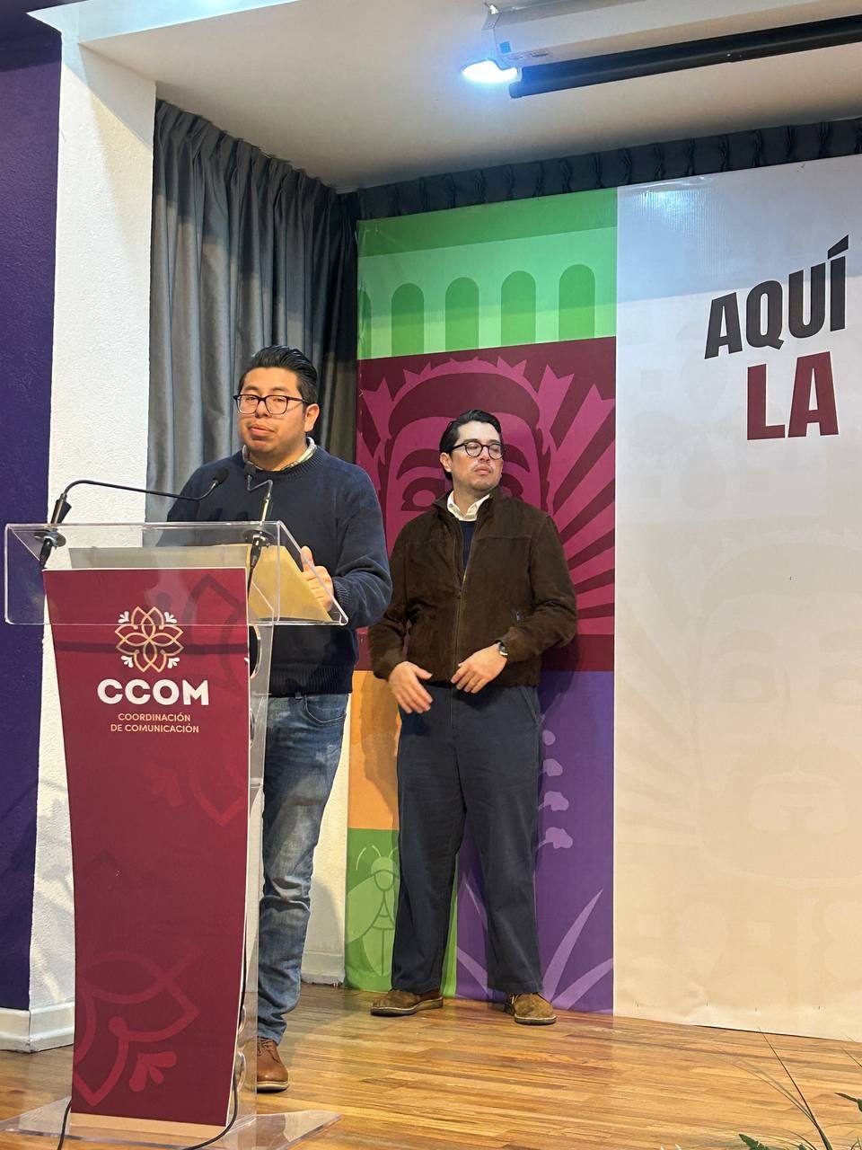 Un hombre habla en un podio de la Coordinación de Comunicación (CCOM) y otro hombre está de pie a su lado