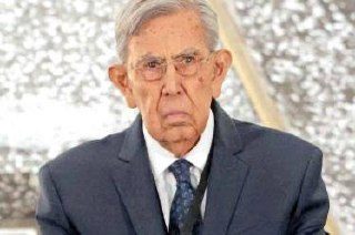 Hombre mayor de cabello canoso y lentes, con traje oscuro y corbata, posa con gesto serio frente a un fondo brillante