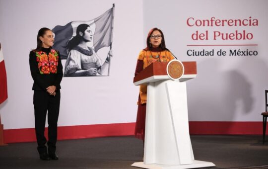 Una mujer habla en un podio en la Conferencia del Pueblo, en Ciudad de México; otra mujer la observa de pie a un lado