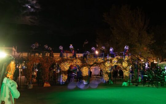 Estructura iluminada con arcos de flores amarillas y figuras con máscaras sobre una plataforma, de noche, en el festival...