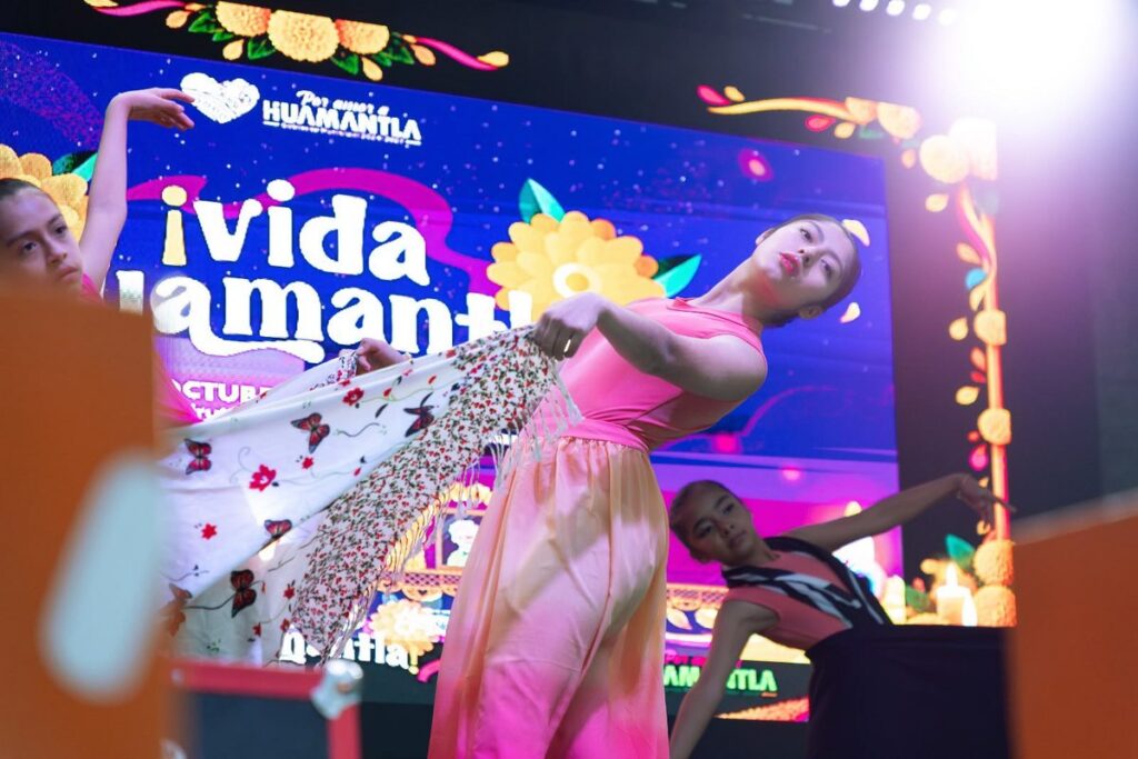 Bailarina con vestido rosa y chal floral baila en el escenario del festival Vida Huamantla 2026, frente a una pantalla...