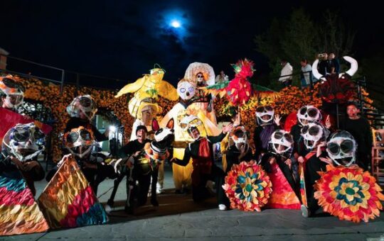 Grupo de artistas con máscaras de calavera y gigantescos títeres coloridos posan de noche frente a muros de cempasúchil...